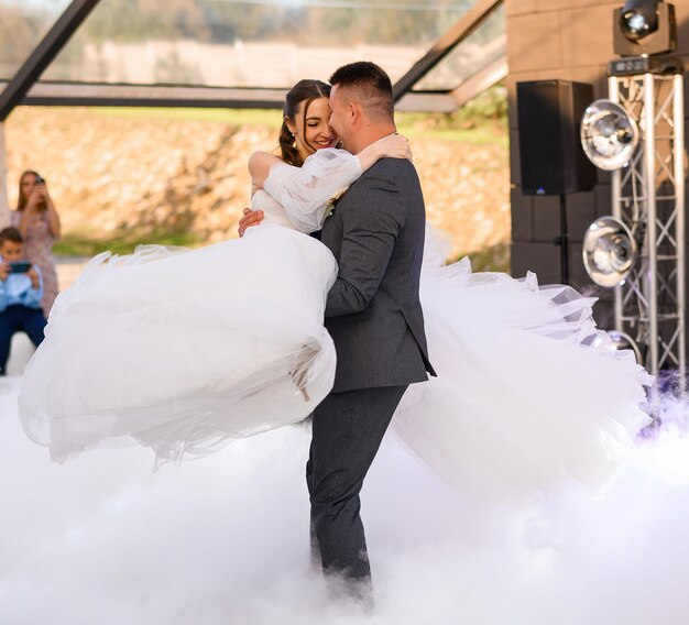 Happy groom holding on hands beautiful bride while dancing and circling on the outdoor terrace in smoke having first wedding dance