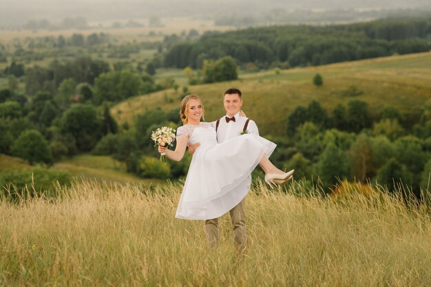Счастливый жених держит на руках свою прекрасную невесту, против красивый пейзаж.
