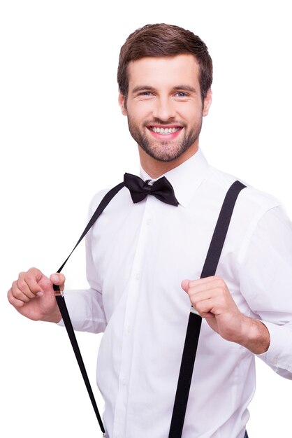 Happy groom. Portrait of handsome young man in white shirt and bow tie adjusting his suspenders and smiling at camera while standing isolated on white