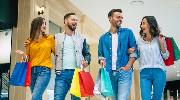 종이 봉투와 함께 캐주얼에 흥분된 아름다운 현대적인 세련된 친구들의 행복한 그룹이 쇼핑하는 동안 쇼핑몰에서 걷고 있습니다.