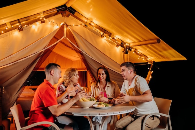 Happy group of friends relaxing in glamping and drinking wine on summer evening near cozy bonfire Luxury camping tent for outdoor recreation and recreation Lifestyle concept