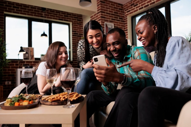 Happy group of multiethnic friends at wine party watching funny clips on mobile phone. Diverse joyful people sitting at home in living room while laughing and celebrating birthday event.