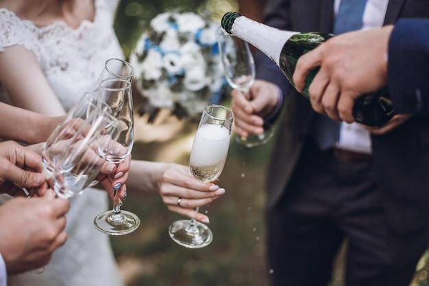 Happy group of people toasting with champagne man holding bottle of champagne and pouring drink into glasses bride bridesmaids and groom groomsmen having fun holiday celebration