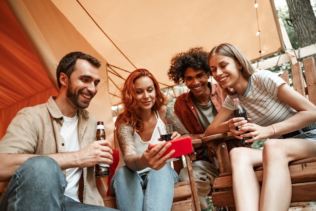 Happy group of young people drinking wine beer using phones social media during picnic in forest smiling millennial users customers talking shopping online sit at table, mobile tech lifestyle concept