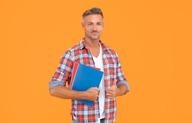 学校の本を握る親指をあげるカジュアルな幸せな男 黄色い背景の教育