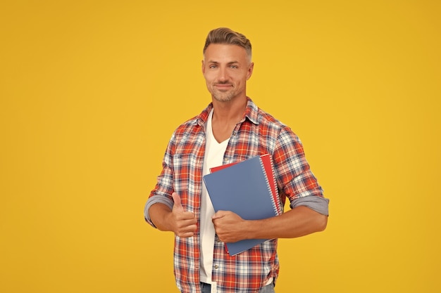 学校の本を握る親指をあげるカジュアルな幸せな男 黄色い背景の教育