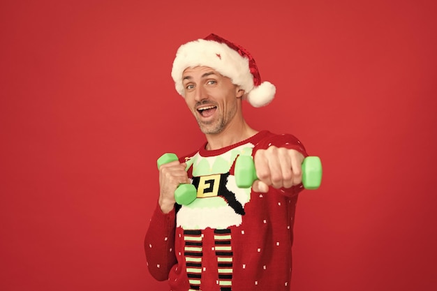 Happy guy in Christmas hat and jumper do dumbell exercises red background Santa
