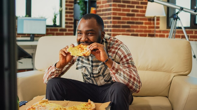 Photo happy guy eating slice of pizza and drinking beer at home, enjoying takeout food and watching television. young man serving takeaway dinner and alcohol, watch movie in living room.