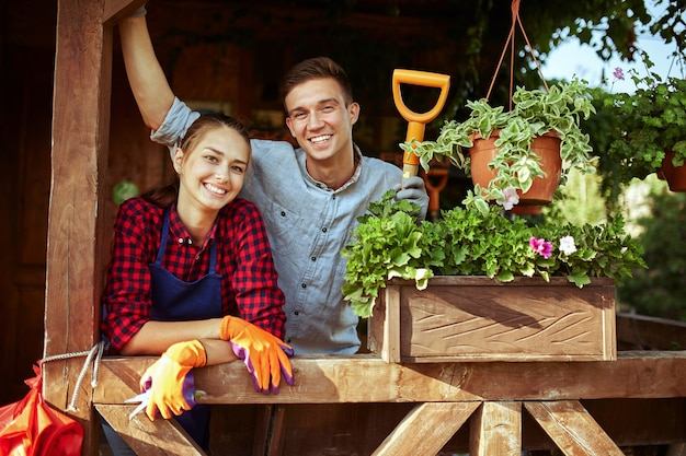 Happy guy and girl gardeners standing on the wooden veranda with plants on a sunny day .