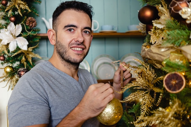 Happy guy holds golden christmas ball and decorates the tree