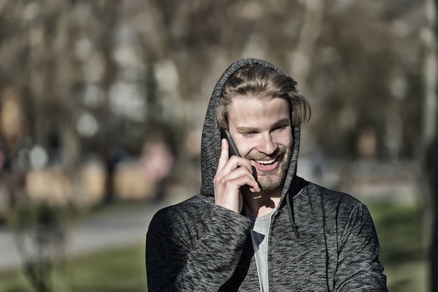 Happy guy in hood talk on mobile phone on sunny outdoor. Bearded man smile in hood on fresh air. Fashion guy smiling with smartphone in casual sweatshirt. New technology, 4G and 3G.