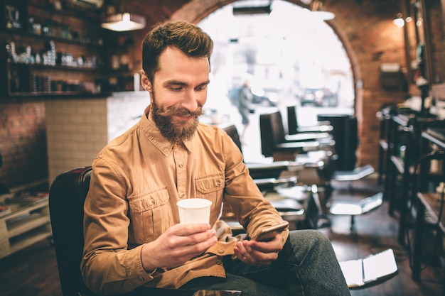 Happy guy is looking at the phone and smiling. He is holding a cup of tea in the right hand and enjoying the moment.