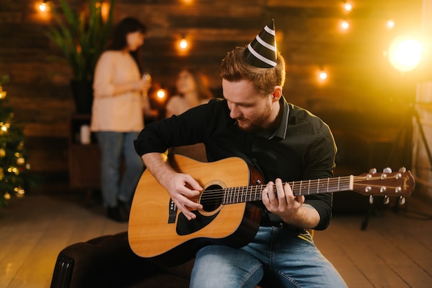 Happy guy is playing guitar. Guy is looking happily and carefree. Man in festive hat celebrating Christmas or new year. Wall with festive illumination in background.