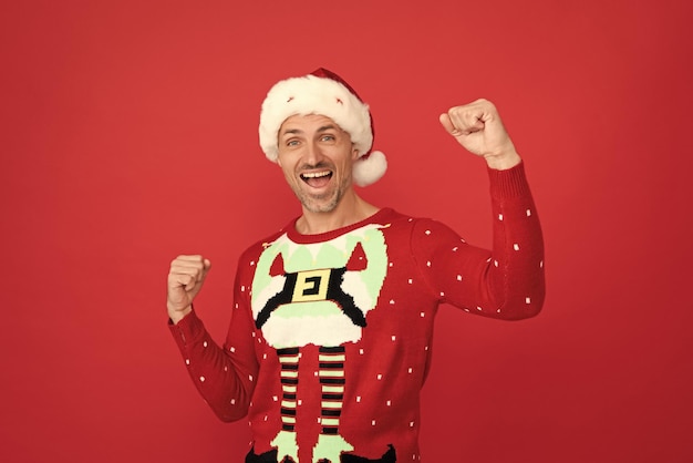 Happy guy in Santa hat and Christmas jumper scream making winning gesture red background new year