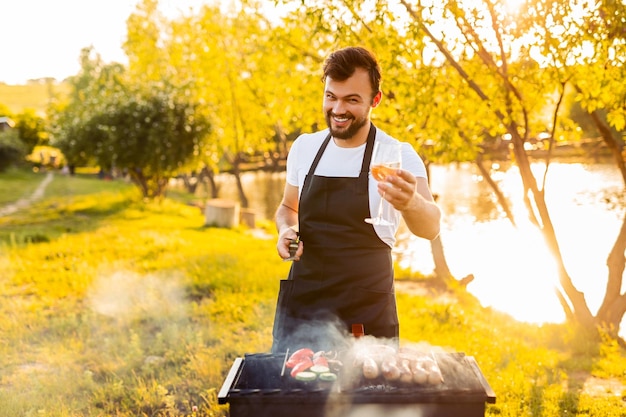 Happy guy toasting to camera while grilling sausages and vegetables in park