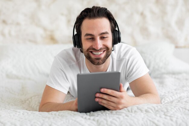 Happy guy using digital tablet and wireless headset bedroom interior