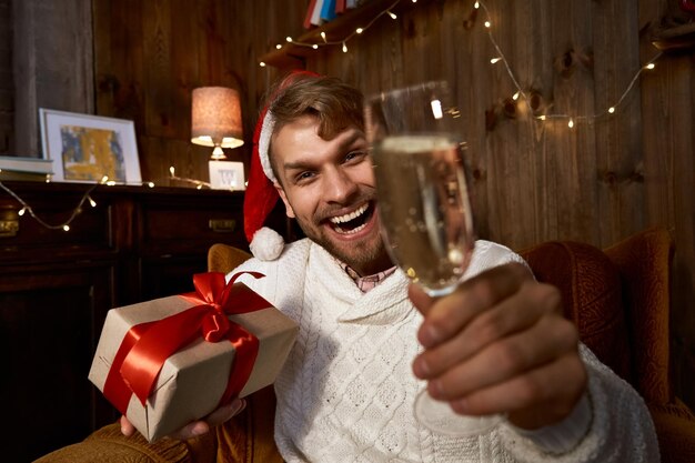 写真 クリスマスのプレゼントとシャンパングラスを持っているサンタの帽子をかぶった幸せな男バーチャルチャットで友人や家族とカメラを見ながらハローと言った新年を祝う遠隔オンラインパーティーウェブカメラビュー