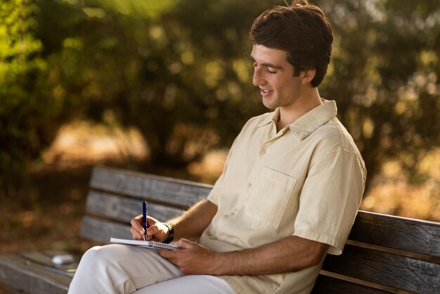 Happy guy writer sitting on bench at park writing notes