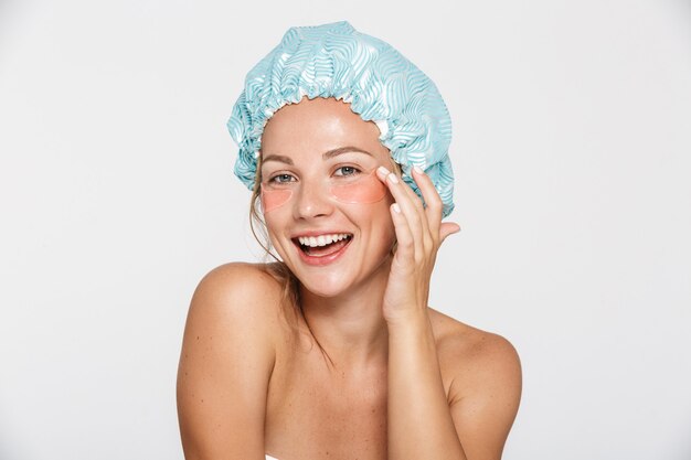 happy half-naked woman wearing cosmetic patches and shower cap smiling isolated over white wall