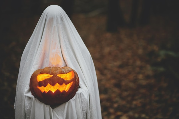 Happy Halloween Boo Spooky ghost holding glowing jack o lantern in moody dark autumn forest Person dressed in white sheet as ghost with pumpkin standing in atmospheric evening woods