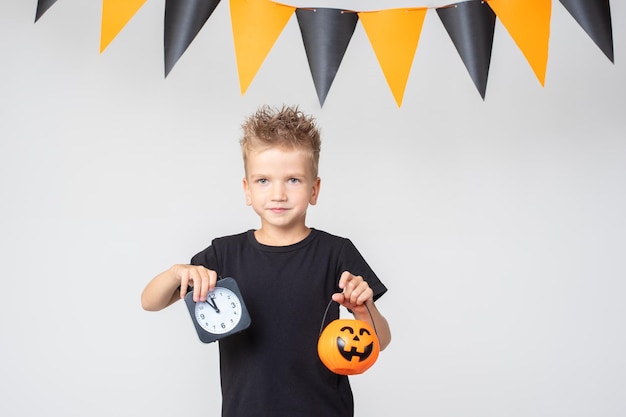 Happy Halloween boy in a black Tshirt holding a pumpkinshaped bucket with treats and a black alarm clock