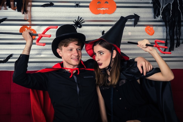 Happy Halloween. couple young caucasian man and woman in vampire and witch clothing holding devil trident and sitting on sofa in halloween party at home, holiday and halloween festival concept