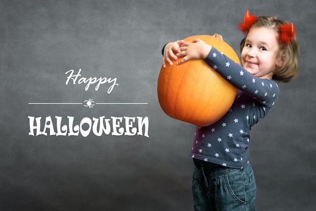 Happy halloween funny little girl holds big pumpkin and smiles adorable child and halloween typography cute toddler with costume horns having fun studio portrait of cheerful kid on halloween