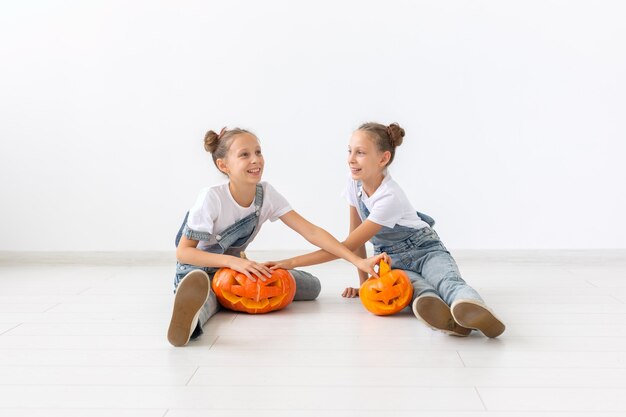 ハッピーハロウィン、休日、子供時代のコンセプト-カボチャのジャック・オー・ランタンとかわいい双子の姉妹の女の子が屋内で時間を過ごしながら楽しんでいます