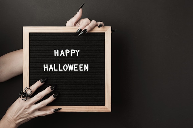 Happy Halloween on letter board in hands with long black nails and spider ring on black background. Halloween holiday concept.