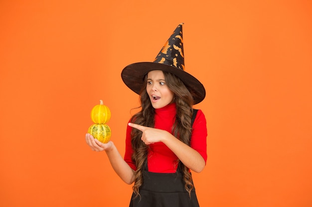 Happy halloween party kid wearing witch cosplay costume and having fun while celebrating autumn traditional holiday pointing finger on pumpkin, halloween jack o lantern.