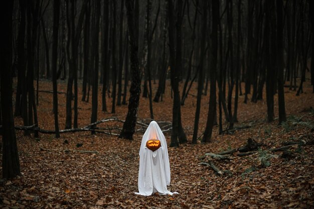 Happy Halloween Spooky ghost holding glowing jack o lantern in moody dark autumn forest Person dressed in white sheet as ghost with pumpkin standing in atmospheric evening woods Boo