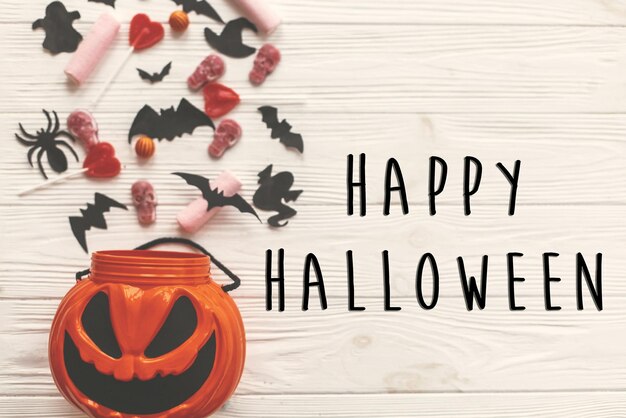 Happy halloween text sign flat lay jack o lantern bucket with holiday candy batsspiders skulls on white rustic wood space for text season's greeting card