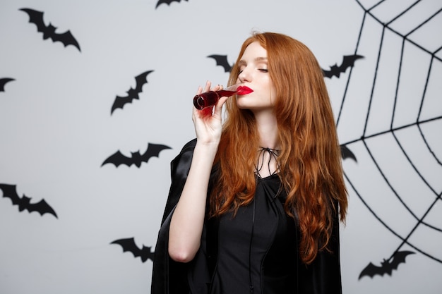 Happy Halloween Witch drinking blood over dark grey studio background with bat and spider web.