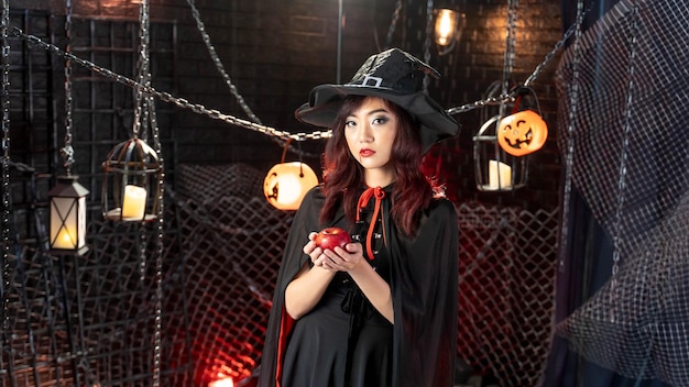 Happy halloween young asian woman in witch costume wearing witch hat holding apple in halloween theme