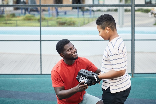 Happy handsome African-American father with beard chatting with teenage son in baseball glove while preparing him for game