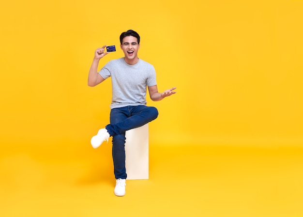 Happy handsome Asian man showing credit card in hand isolated on yellow background.