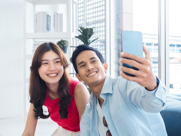 Happy handsome Asian man and young beautiful woman selfie while sitting near the glass window at white living room in high building Cheerful couple taking portrait selfie with smart mobile phone