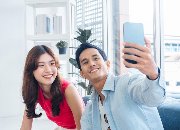 Happy handsome Asian man and young beautiful woman selfie while sitting near the glass window at white living room in high building Cheerful couple taking portrait selfie with smart mobile phone