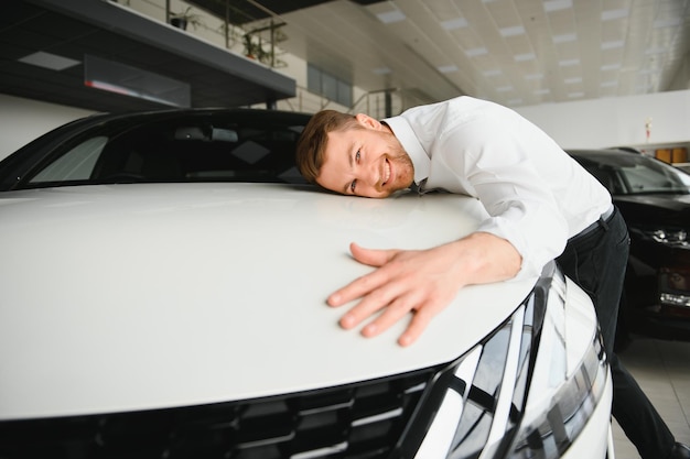 Happy handsome bearded man buying a car in dealership guy hugging hood of new car