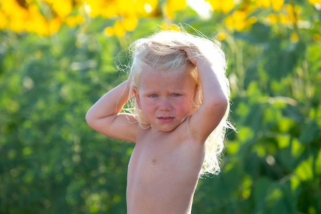 Happy handsome boy with long blond hair stands in a field of blooming sunflowers