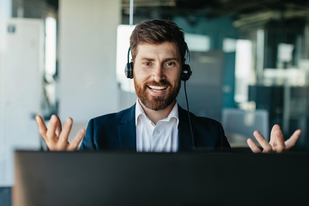 Happy handsome businessman in headset working on laptop and gesturing to webcam having video call in office interior