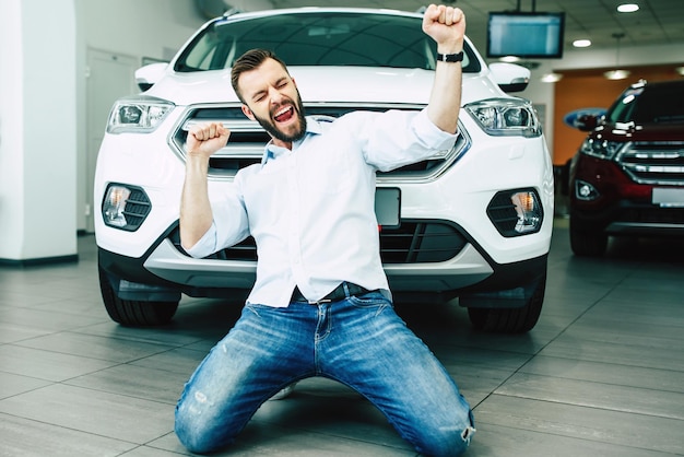 Happy handsome man buying a new white car in dealership and dancing on the floor against the auto