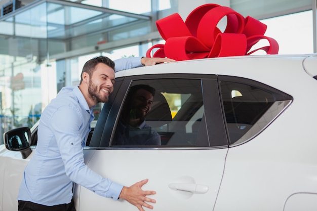 ディーラーで彼の新しい車を抱きしめる幸せなハンサムな男。屋根の上の赤い弓が付いているギフト車を抱いて興奮している男性ドライバー