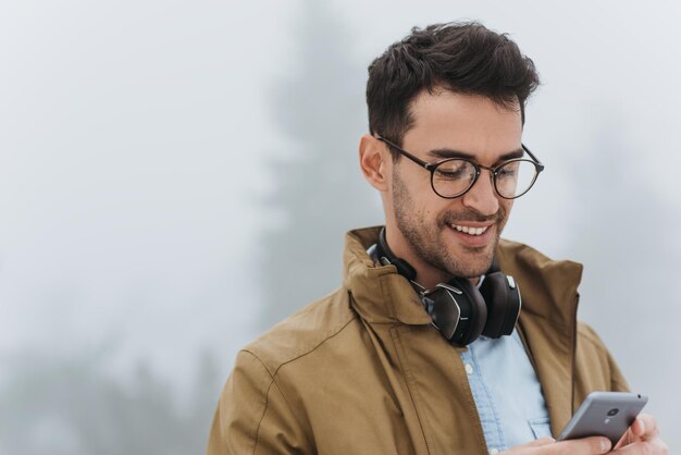 Happy handsome man looking down wearing eyeglasses coat and black headphones to listening the music from smartphone in the misty park outdoor People technology and lifestyle concept