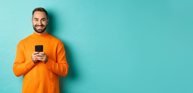 Foto uomo bello felice che scrive messaggio sul telefono cellulare che tiene smartphone e sorridente in piedi contro t