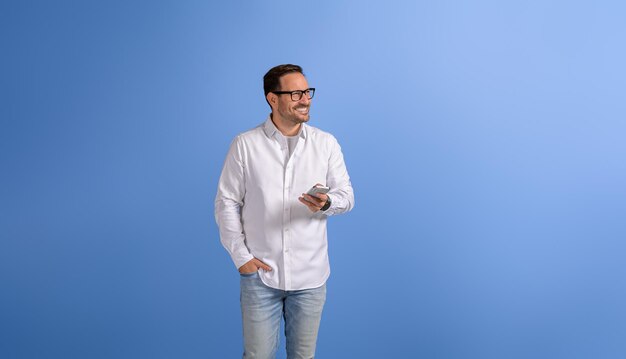 Happy handsome manager with hand in pocket using smart phone and looking away over blue background