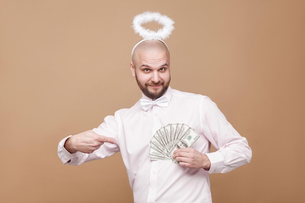 Happy handsome middle aged bald angel in shirt and white halo standing, holding and pointing at many dollars and looking at camera with smile. indoor studio shot, isolated on light brown background.