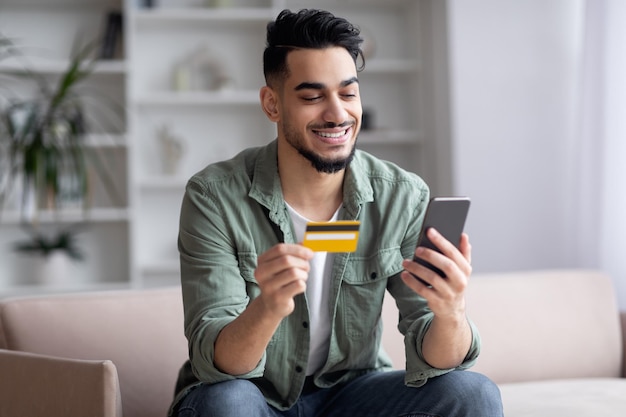 Happy handsome middle eastern guy shopping online with smartphone and credit card