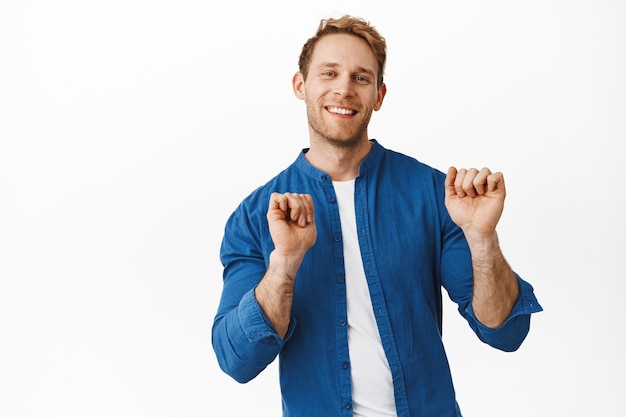 Happy handsome redhead man smiling, looking at front joyful and dancing, raising hands up, moving clumsy but cute on dance floor, standing over white wall