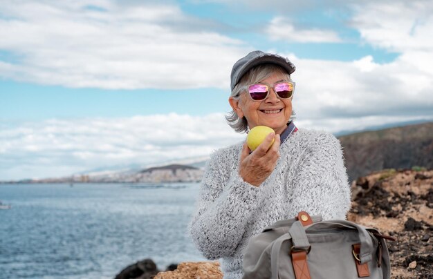 Happy handsome senior woman sitting by the sea eating an apple enjoying freedom vacation